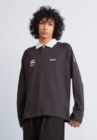 Sort langærmet polo skjorte med hvid krave, har et rundt logo og tekst på brystet, med en afslappet pasform og blød tekstur.