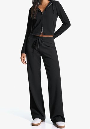 Femme portant un sweat à capuche noir court avec fermeture éclair et un pantalon large assorti à cordon de serrage, associé à des baskets blanches, debout avec la main sur la hanche.