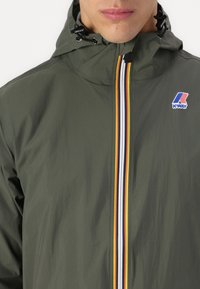 Chaqueta impermeable verde con capucha, que presenta un cierre frontal y rayas verticales multicolores. Logo en el lado izquierdo del pecho. Textura suave.