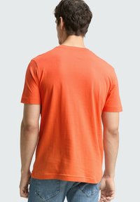 Pomarańczowy t-shirt z krótkim rękawem i okrągłym dekoltem, o gładkiej fakturze, noszony z niebieskimi jeansami. Widok od tyłu ukazuje prosty wzór, bez grafik.