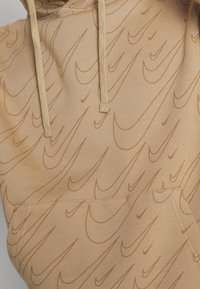 Beige huvtröja med ett mönster av upprepade Nike-swooshar i en något mörkare nyans. Har en framficka och justerbara dragskor.