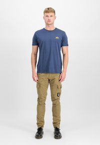 Navyblaues Baumwoll-T-Shirt mit kurzen Ärmeln und einem kleinen gelben Logo. Khaki-Cargohosen mit mehreren Taschen und einem Aufnäher am Oberschenkel. Schwarze Schuhe.