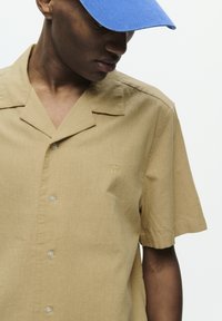 Chemise à manches courtes, beige, avec un motif subtil et un petit logo brodé. Elle présente un col classique et des boutons transparents.