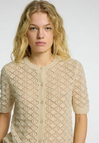 Beige pletený cardigan s krátkými rukávy, se vzorem diamantů, texturovaným povrchem a předním zapínáním na sedm knoflíků.