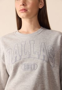 Šedá mikina s vyvýšeným nápisem "DALLAS" a "1967." Okruglý výstřih a jemná textura, s žebrovanými manžetami a spodním lemem.