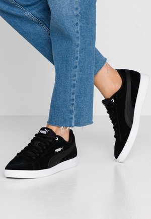 Nogi w niebieskich dżinsach i czarnych butach sportowych Puma z białymi podeszwami, jedna noga lekko uniesiona na jednolitym szarym tle.