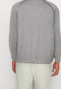 Grå sweatshirt med långa ärmar, mjuk textur och lätt samling i nederkant. Har svarta accentsömmar längs axlarna.