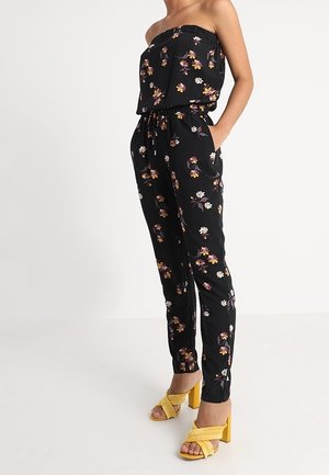 Kvinde iført sort blomstret skulderfri jumpsuit med lommer og gule højhælede sandaler, stående med den ene hånd i lommen.