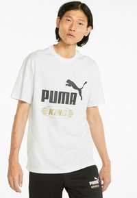 Bílá bavlněná tričko s logem černého skákajícího pantera a výrazným textem "PUMA KING" v černé a zlaté barvě. Krátké rukávy a design s kulatým výstřihem.
