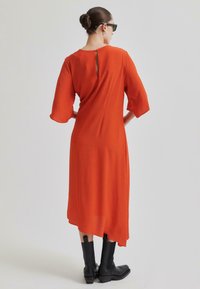 Oranžinė midi suknelė su pusiau ilgomis rankovėmis, turinti atvirą sprandą. Dėvima su juodomis kulkšnies botas, ant neutralaus fono.