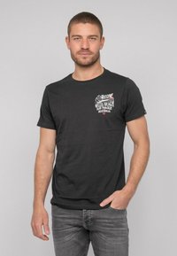 T-shirt en coton noir à manches courtes, col rond, avec un imprimé graphique blanc et rouge sur le côté supérieur gauche. Porté avec un jeans gris.
