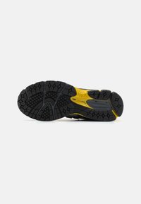 Semelle de chaussure de sport noire et jaune avec un motif de bande de roulement texturée sur fond blanc, mettant en valeur l'adhérence et la durabilité.