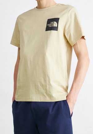 T-shirt en coton beige avec un col rond, des manches courtes et un patch logo noir sur la poitrine gauche. Le tissu est lisse et léger.