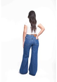 Pantalones vaqueros acampanados de tiro alto en denim azul medio con una textura suave. Cuenta con dos bolsillos traseros y un parche de cuero marrón con la marca en la cintura.
