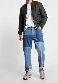 Svart bomberjacka med dragkedjefickor, vit t-shirt och pippjeans i olika blå nyanser. Grå sneakers med röda accenter.