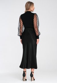 Czarna sukienka z przezroczystymi, bufiastymi rękawami z czarnymi kropkami. Gładka satynowa spódnica, do kolan, o dopasowanym kroju oraz sandały na kostkę.
