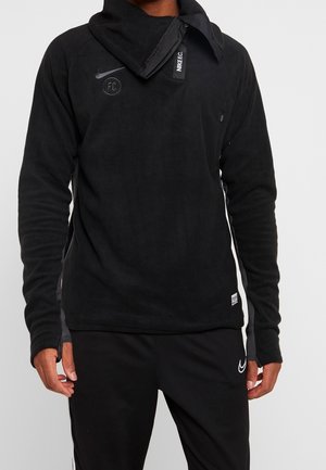 Mies, joka pukeutunut mustaan Nike FC -fleecetoppaan, jossa on korkea kaulus, ja mustiin Nike-verkkareihin, seisomassa yksinkertaista taustaa vasten.