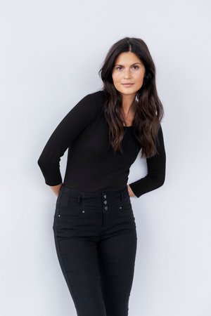 Donna con capelli lunghi e scuri, indossa un top nero a maniche lunghe e jeans neri a vita alta, in piedi contro una parete chiara, mani dietro la schiena.