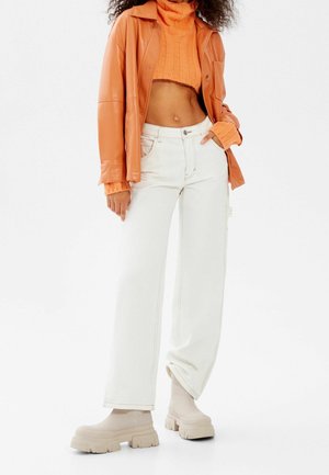 Femme portant un pull court en maille orange, une veste en cuir orange, un pantalon blanc à jambes larges et des bottes beige à semelle épaisse, debout devant un fond blanc.