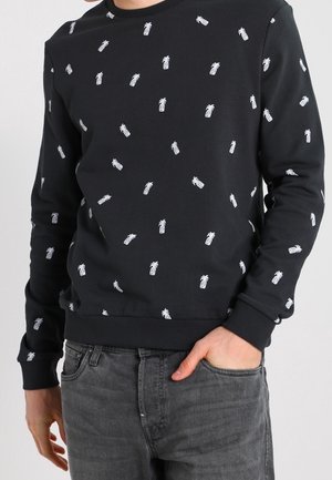 Mann trägt schwarzen Pullover mit kleinen weißen Ananasmustern, kombiniert mit dunkelgrauen Jeans, eine Hand in der Tasche.