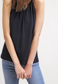 Blusa sin mangas de color negro con escote fruncido, hecha de una tela ligera. Combinada con jeans de color azul claro, que muestran un ajuste relajado.