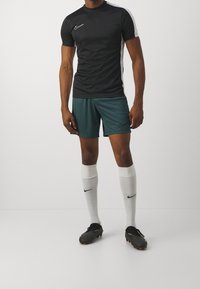 Camisa negra de manga corta de Nike con acentos blancos, pantalones cortos verdes, calcetines blancos y botines negros. El atuendo está diseñado para deportes.