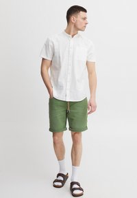 Camicia bianca a maniche corte con colletto e taschino frontale, abbinata a pantaloncini verdi risvoltati e sandali marroni, con calzini bianchi visibili.