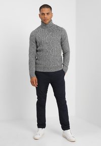 Suéter gris de punto con un cuello alto, textura gruesa y puños acanalados, combinado con pantalones oscuros y zapatillas blancas. Fondo sencillo.