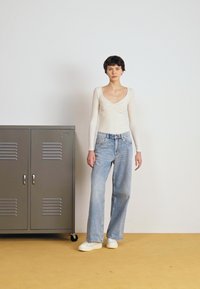 Maglietta beige a maniche lunghe con scollo a V, abbinata a jeans a gamba larga di colore blu chiaro. Sneakers bianche. Armadietto metallico grigio sullo sfondo.