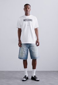 Balts Moschino T-krekls ar melnu logo, apvienots ar zilām džinsa šortiem ar izbalējušu apdari un nodiluma akcentiem. Melni apavi un balti zeķes.