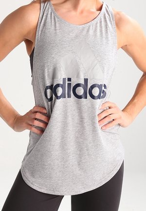 Vrouw draagt een grijs mouwloos Adidas sporttop met centraal logo, handen in de zij en zwarte legging.