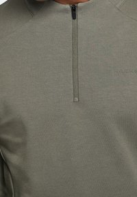 Sudadera verde con cremallera en una suave tela texturizada. Presenta un cuello alto y un sutil logotipo a lo largo de la costura lateral. Diseño minimalista.