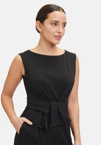 Black sleeveless jumpsuit with a fitted bodice and a textured bow accent at the waist. Features side pockets and a smooth fabric finish.