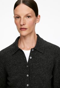 Dunkelgrauer Wollcardigan mit kragen und Knopfverschluss sowie subtiler Textur, über einem weißen Oberteil getragen.