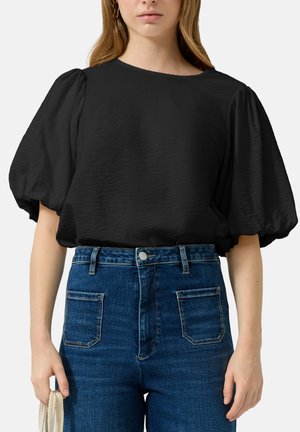 Kobieta w czarnej bluzce z bufiastymi krótkimi rękawami włożonej w niebieskie, jeansowe spodnie z wysokim stanem, trzymająca małą beżową torebkę przy boku.