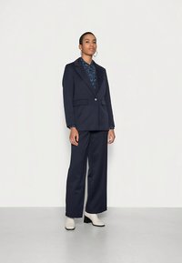 Blazer azul-marinho ajustado com dois bolsos frontais, combinado com calças largas de perna larga combinando. Por baixo, uma blusa azul com padrão. Botas brancas até ao tornozelo.