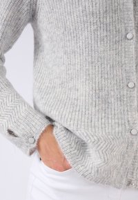 Graue Strickjacke mit strukturiertem Fischgrätmuster, mit weißen Knöpfen und lockerem Sitz. Weiches, gemütliches Material, lange Ärmel.