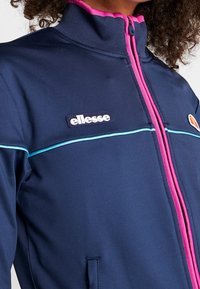 Marinblå zip-up jacka med hög krage, rosa detaljer och ljusblå kant. Har en logotypmärke och texturerat tyg. Två sidofickor.