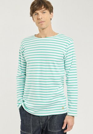 Homme portant un t-shirt à manches longues rayé horizontalement vert et blanc avec un jean foncé, debout devant un fond clair uni.