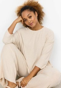 Beige ribbat loungedress med bred, rund halsringning och lösa, långa ärmar. Modell som bär ljusa skor, sitter med handen på hakan.