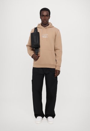 Felpa beige con tasca frontale e logo, abbinata a pantaloni cargo neri larghi. Il modello tiene una borsa a tracolla nera e indossa sneaker bianche.