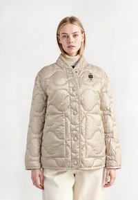 Beige gewatteerde pufferjack met een hoge kraag, drukknoopsluiting aan de voorkant en een golfpatroon textuur. Gestyled met een lichte coltrui eronder.