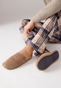 Braune gewebte Hausschuhe mit strukturiertem Obermaterial, Gummisohle und dezentem Logodetail, kombiniert mit karierten, beige-blauen Pyjamahosen.