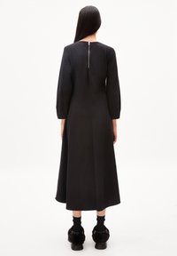 Zwarte, enkellange jurk met lange mouwen, voorzien van een ronde hals en een rits aan de achterkant. Glad materiaal en een eenvoudig ontwerp. Zwarte schoenen met bontaccenten.