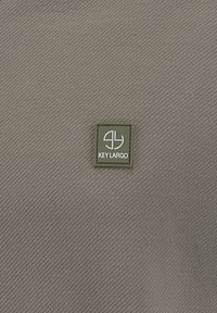 Klein olijfgroen vierkant patch met wit abstract logo en tekst "KEY LARGO" op een gestructureerde grijze stoffen achtergrond.