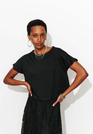 Femme aux cheveux courts et bouclés portant des boucles d'oreilles créoles, des colliers en argent superposés, un haut noir et une jupe en dentelle noire, posant les mains sur les hanches.