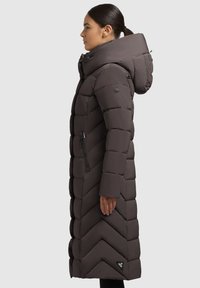 Donna di profilo che indossa un lungo cappotto invernale trapuntato di colore marrone scuro con colletto alto, cappuccio, tasche laterali con zip e un motivo a chevron.