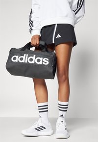 Svart duffelväska med vit "adidas"-text, robusta handtag och textilstruktur. Bärs med svarta shorts och vita sportstrumpor med ränder.