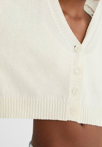 Cream-colored cropped cardigan featuring a V-neck, ribbed hem, and four large buttons made of plastic. Soft, textured knit fabric.