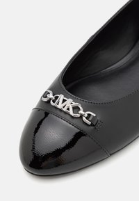 Zwarte leren platte schoen met een glanzende punt. Heeft een zilveren kettingaccent met het "MK"-logo op de wreef. Glad textuur, minimalistisch ontwerp.